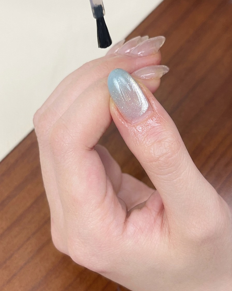 연한 블루 쉬머 캣아이 젤 네일 위에 투명한 광택 탑 코트를 브러쉬로 바르는 모습.