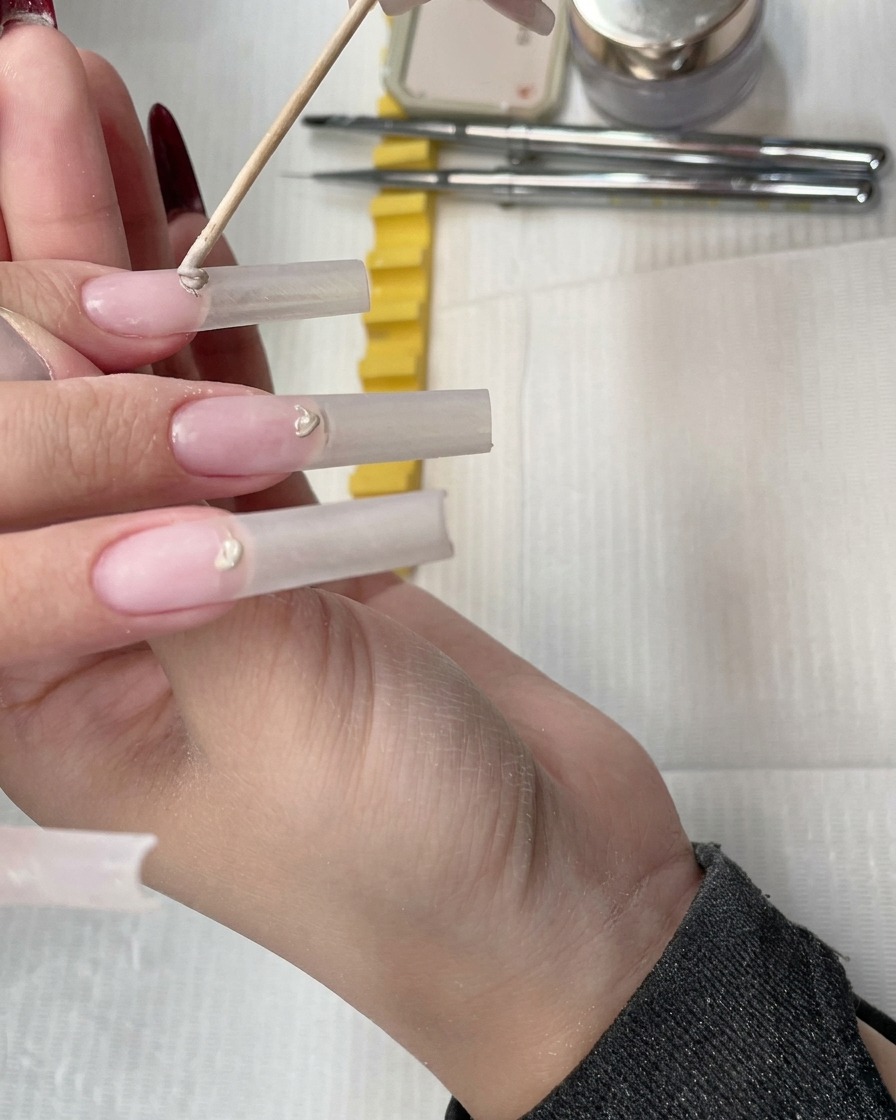 우드 스틱을 사용하여 투명 연장 네일의 큐티클 라인에 누드 컨실러 젤을 작게 올리는 모습.