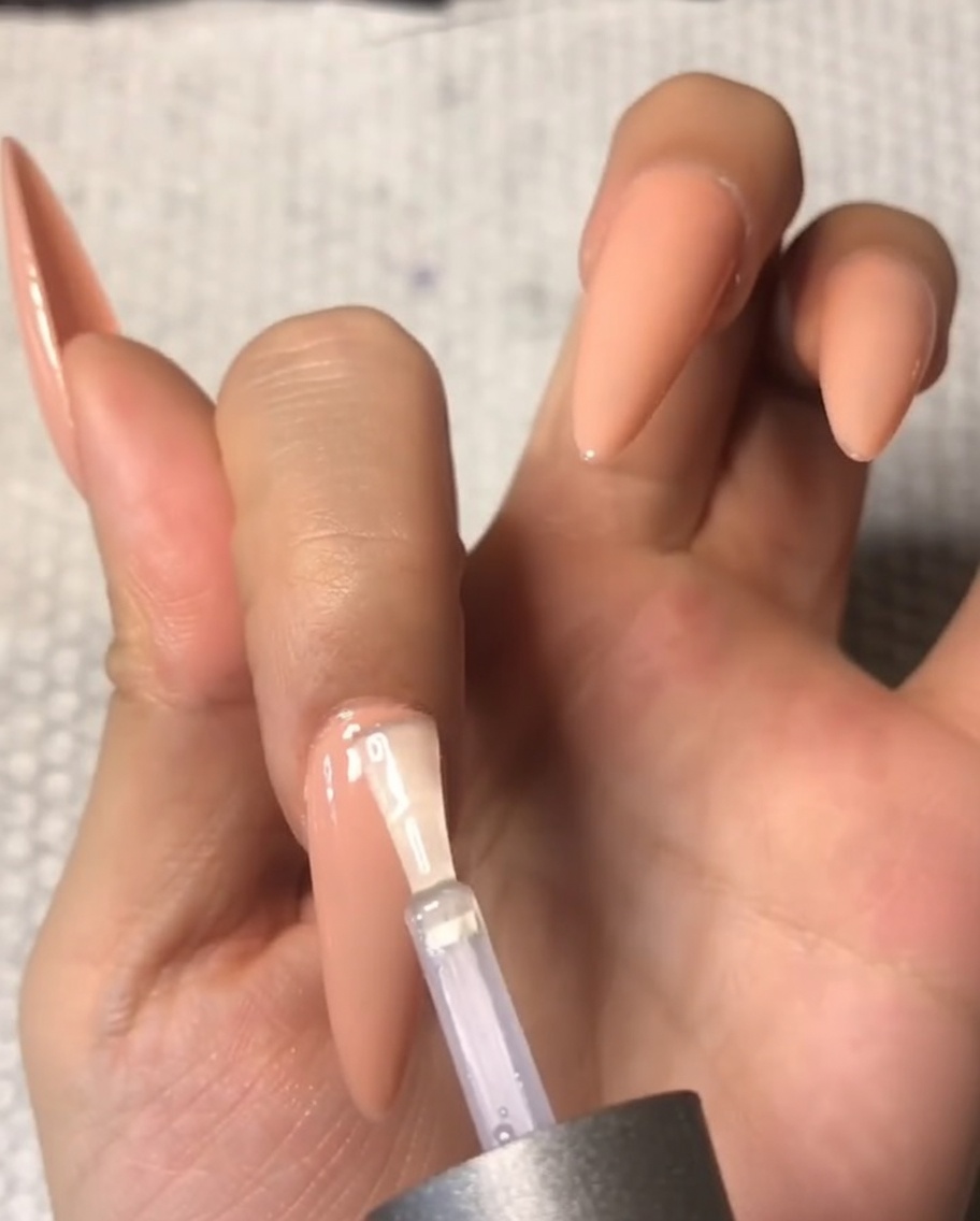 누드톤 아몬드형 중지 손톱 큐티클 라인에 템퍼드 탑코트를 조심스럽게 바르는 브러시.