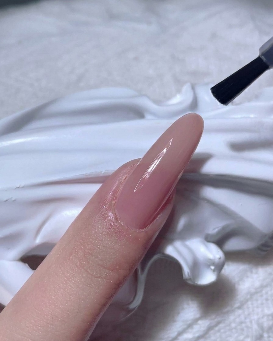 누드 컬러 네일 위에 투명하고 광택 나는 탑코트를 브러시로 바르는 모습.