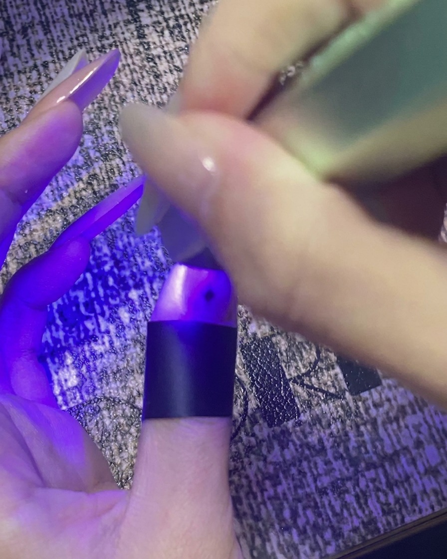 별 네일 아트가 완성된 엄지손가락을 보라색 불빛이 나오는 미니 UV/LED 램프 아래에서 큐어링하는 모습.
