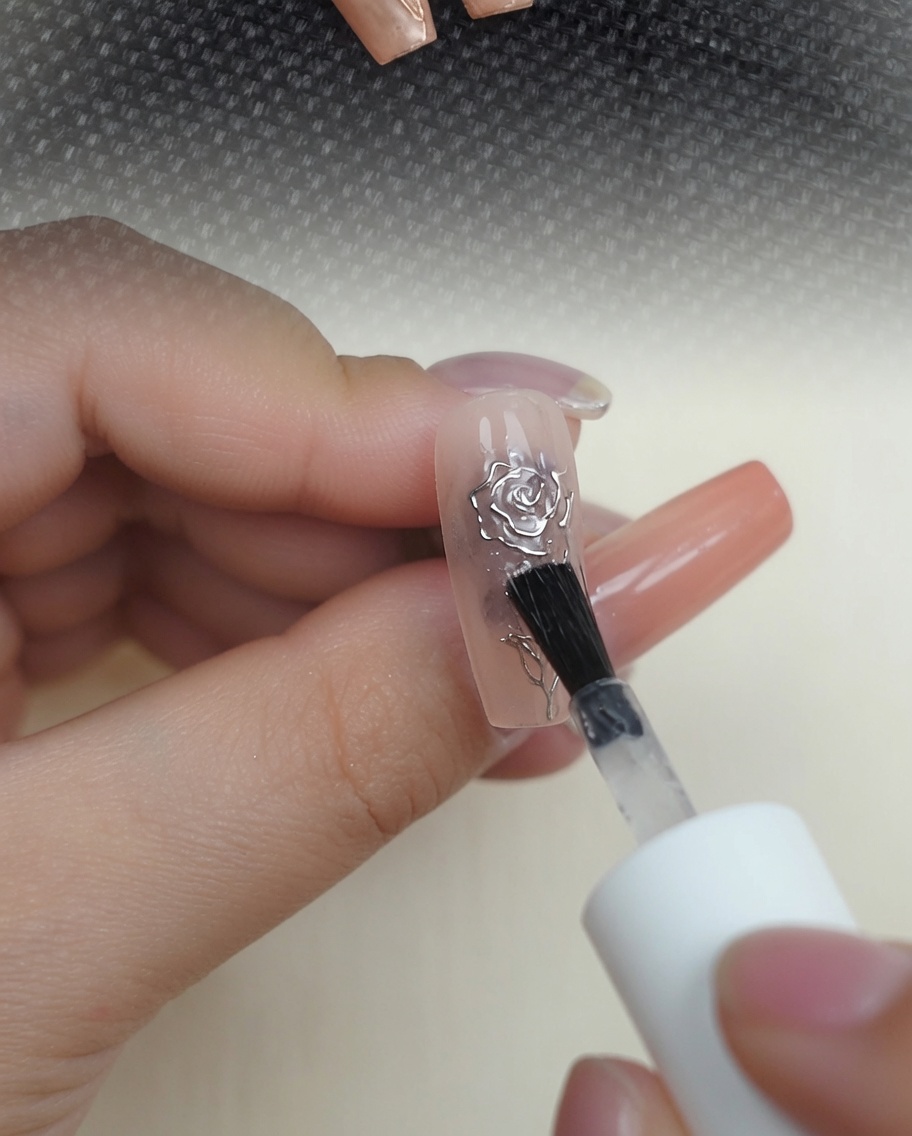 누드 네일 위의 메탈릭 크롬 3D 장미 위에 투명한 글로시 탑코트를 바르는 모습.