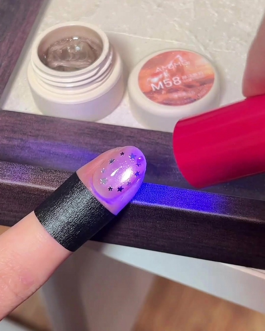 별 글리터 아트를 한 손가락이 UV/LED 램프의 보라색 불빛 아래서 큐어링되는 모습.