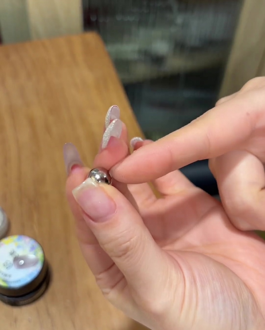 경화되지 않은 반짝이는 캣아이 젤이 발린 손톱 가까이에 작은 구형 은색 자석을 대고 있는 모습.