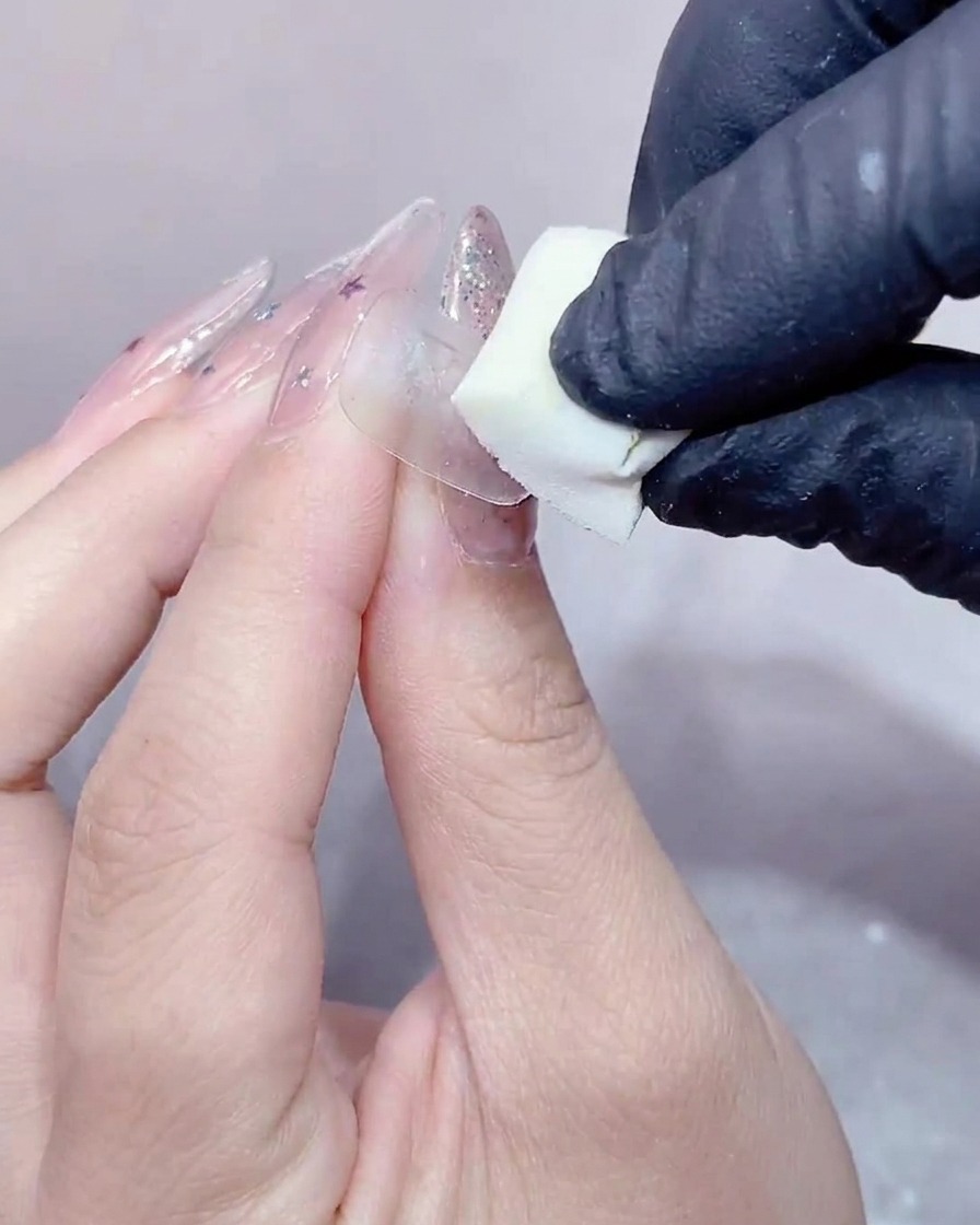장갑을 낀 손으로 투명 젤이 발린 네일 표면을 작은 흰색 메이크업 스펀지로 부드럽게 누르는 모습.