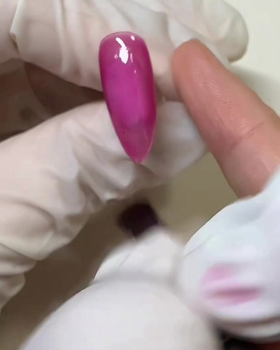 장갑을 낀 손가락이 경화된 핑크 네일 표면 위에 투명한 솔리드 글루를 얇게 펴 바르는 클로즈업 샷.