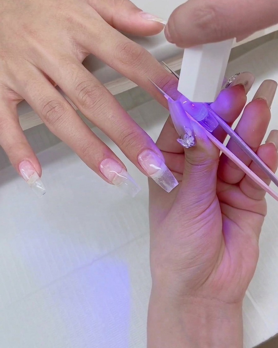 핀셋으로 팁을 고정한 채 투명 네일 연장 팁 위에 미니 UV 플래시 램프를 비추고 있는 손의 클로즈업 샷.