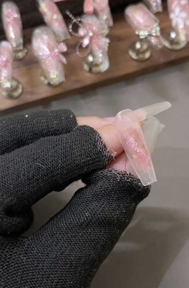반짝이는 핑크빛 샤터드 다이아몬드 글리터 젤이 코팅된 투명한 네일을 장갑 낀 손으로 잡고 있는 모습.