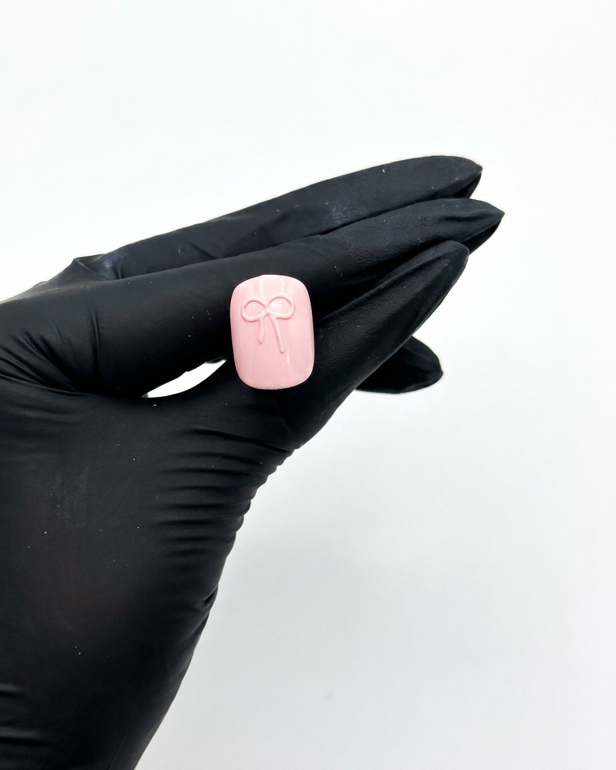 장갑을 낀 손이 3D 리본 디자인이 된 핑크색 압착식 네일을 UV 네일 램프 아래에 대고 경화시키는 모습.