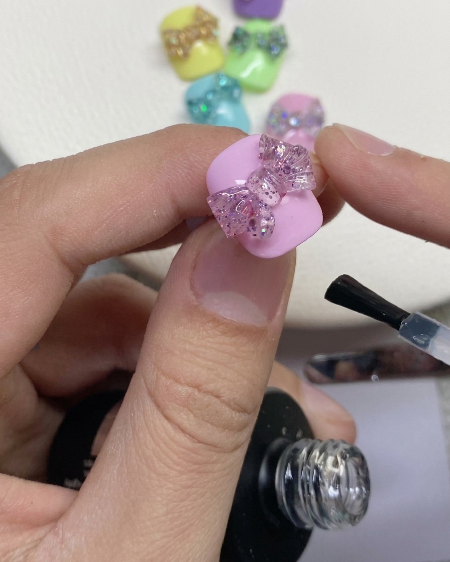 핑크색으로 칠해진 네일 위 3D 글리터 리본 주변에 투명한 탑 코트를 바르고 있는 네일 브러시의 클로즈업.