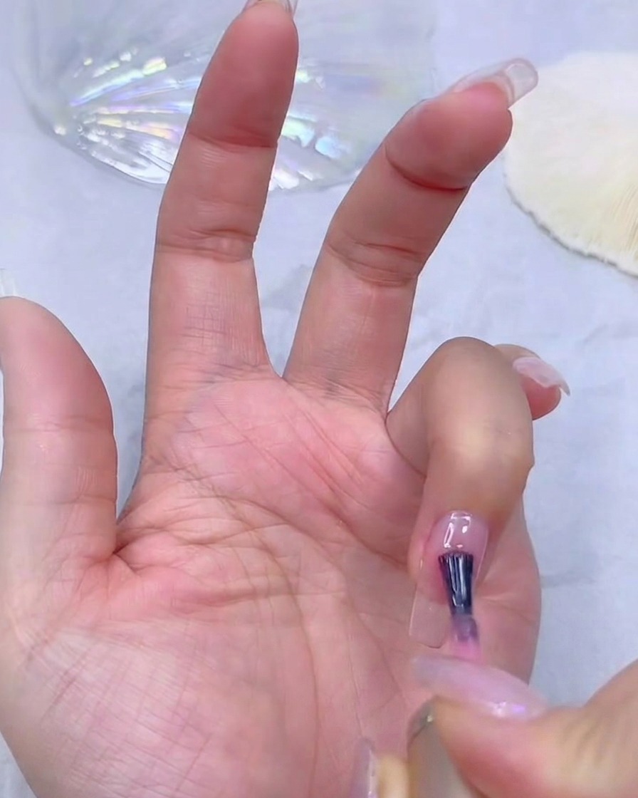 네일 테크니션의 손이 투명한 네일 연장 팁 위에 시어 핑크 젤 베이스코트를 바르고 있는 모습.