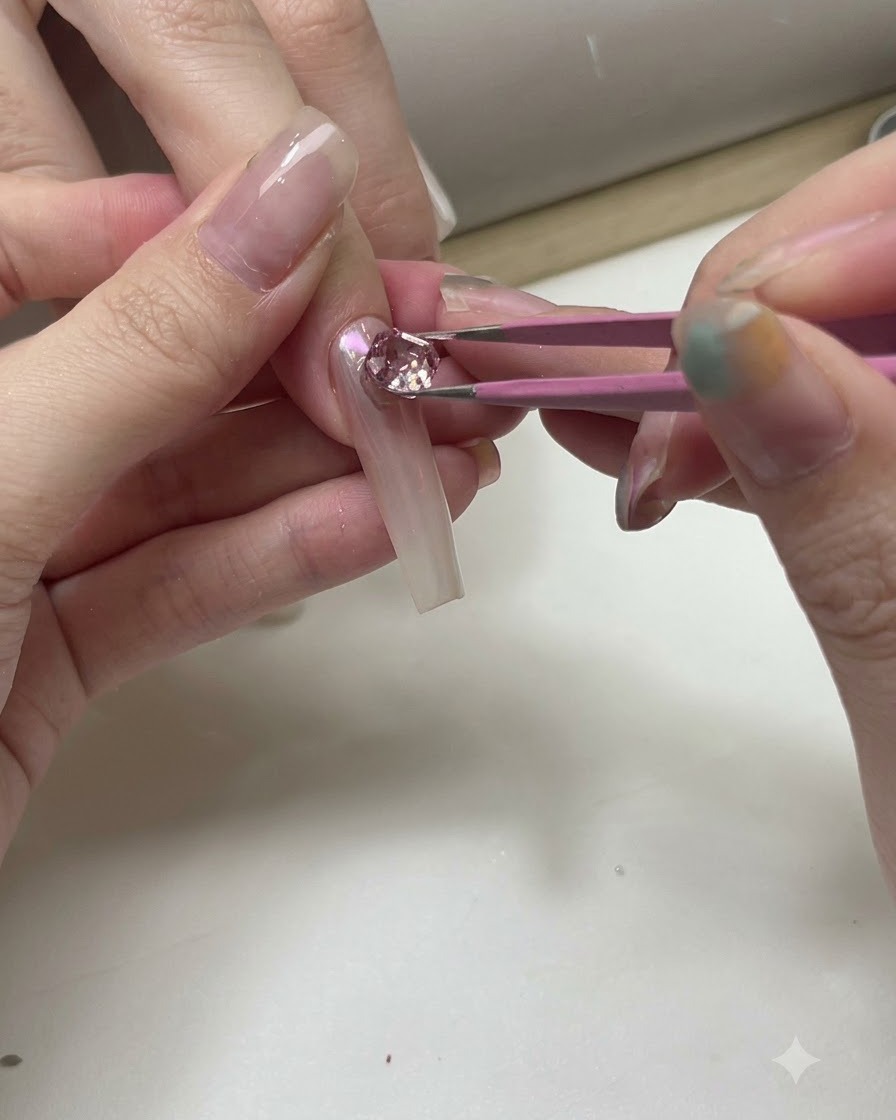 투명한 연습용 네일 팁 위에 핀셋으로 반짝이는 핑크와 클리어 크리스탈을 배치하는 모습.