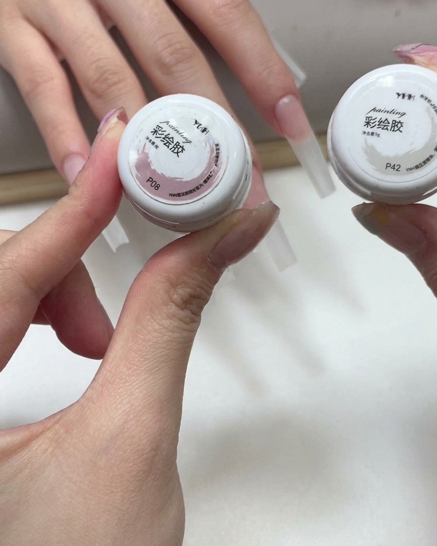배경에 연장된 네일이 보이는 상태에서 핑크와 화이트 컬러 페인팅 젤이 담긴 작은 용기를 들고 있는 손.