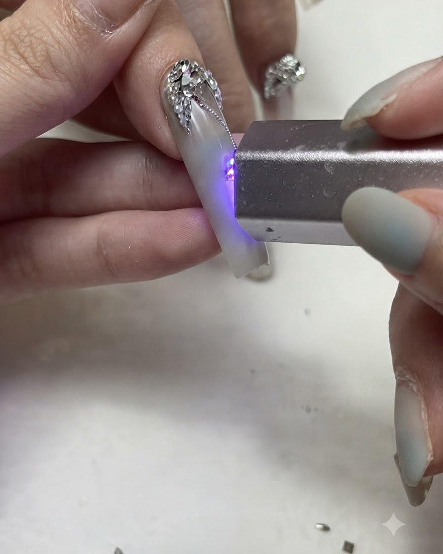 휴대용 UV/LED 플래시라이트로 장식된 네일의 섬세한 실버 젤 라인과 스톤 디테일을 경화하는 모습.