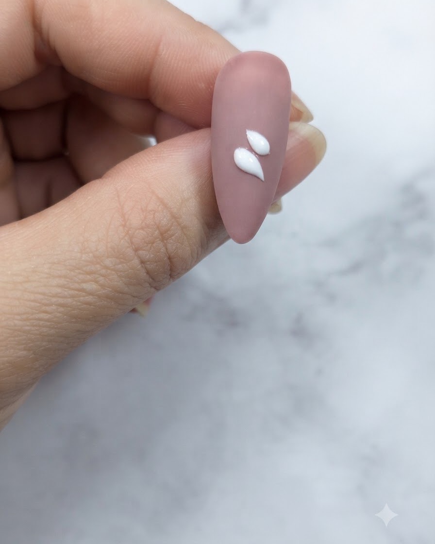 중앙 근처에 적용된 두 개의 선명한 화이트 물방울 모양 꽃잎 스트로크를 보여주는 핑크빛 누드 네일의 클로즈업.