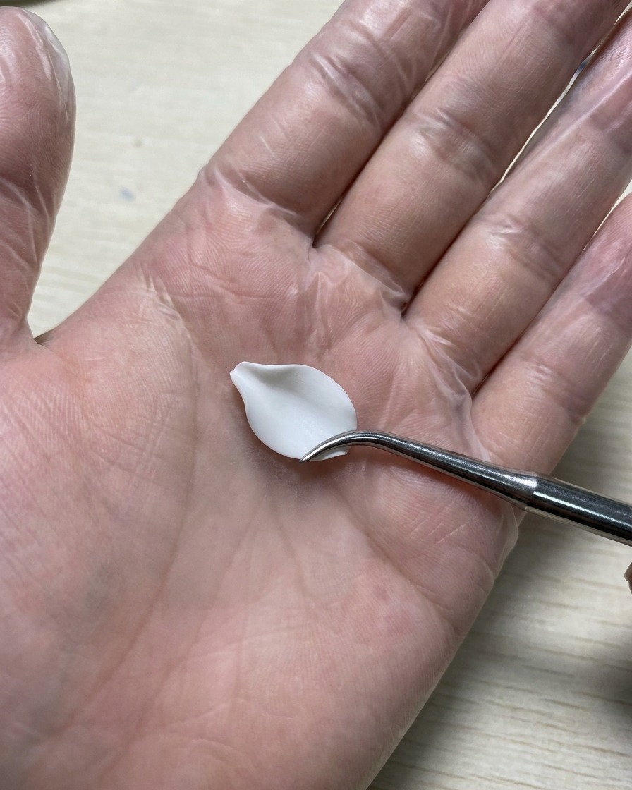 장갑을 낀 손이 실리콘 매트에 놓인 흰색 3D 젤 꽃잎을 매끄럽게 다듬는 실리콘 카빙 툴을 잡고 있는 모습.