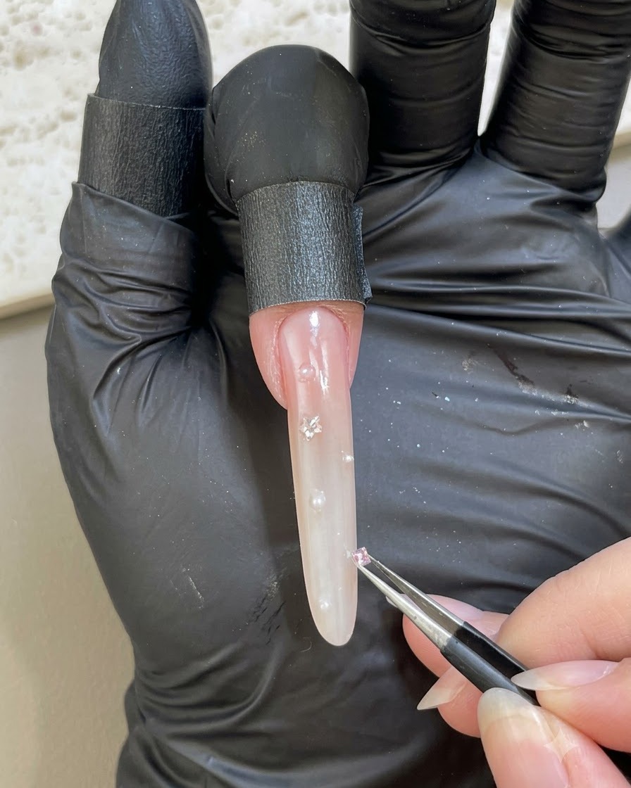 장갑을 낀 손이 핀셋을 사용하여 장식된 네일 표면에 작은 핑크색 스라인스톤을 배치하는 모습.