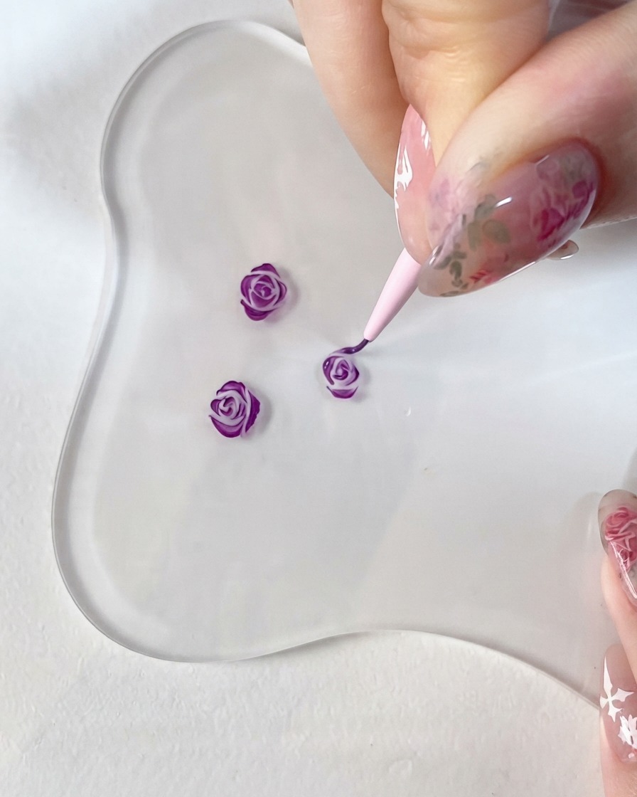 투명 유리 팔레트 위에 놓인 작은 3D 장미 디자인에 세필붓으로 짙은 보라색 포인트를 꼼꼼하게 그리는 클로즈업.