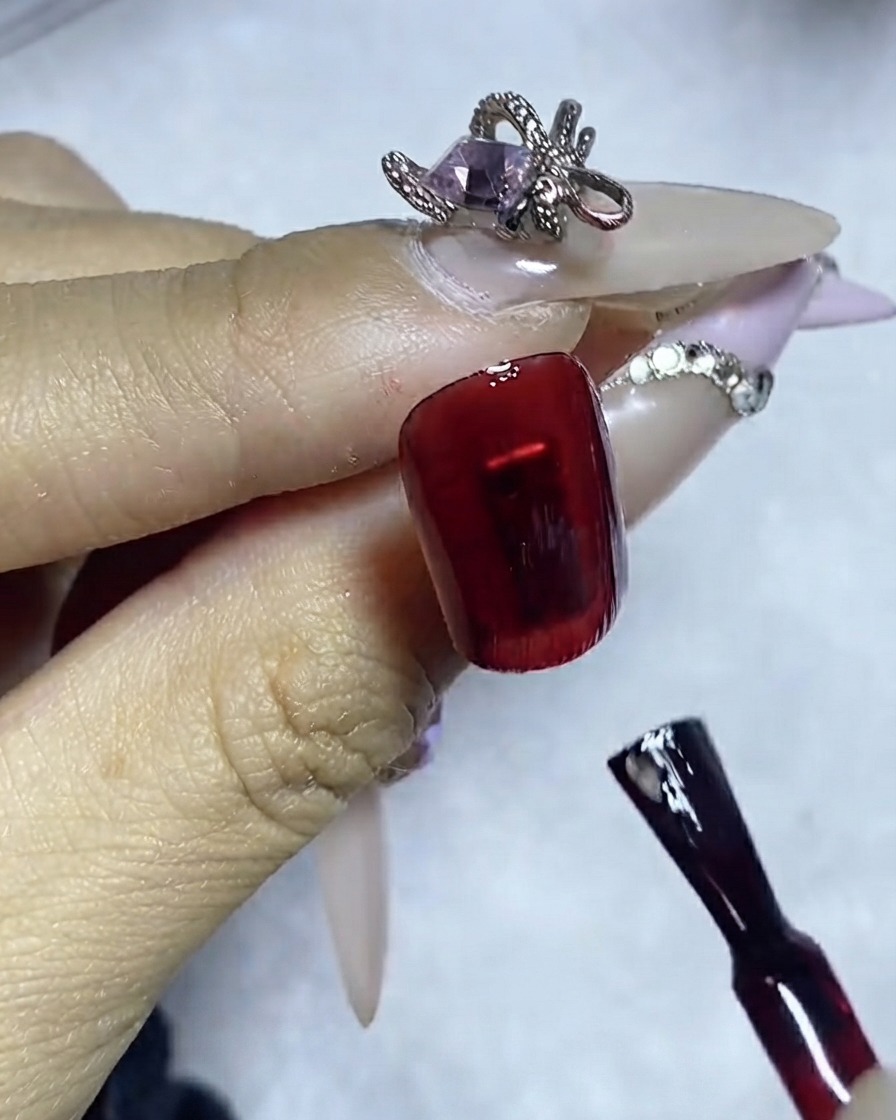 다크 레드 컬러의 폴리시가 발린 네일 표면에 라이너 브러시로 젬 글루를 정교하게 바르는 모습.