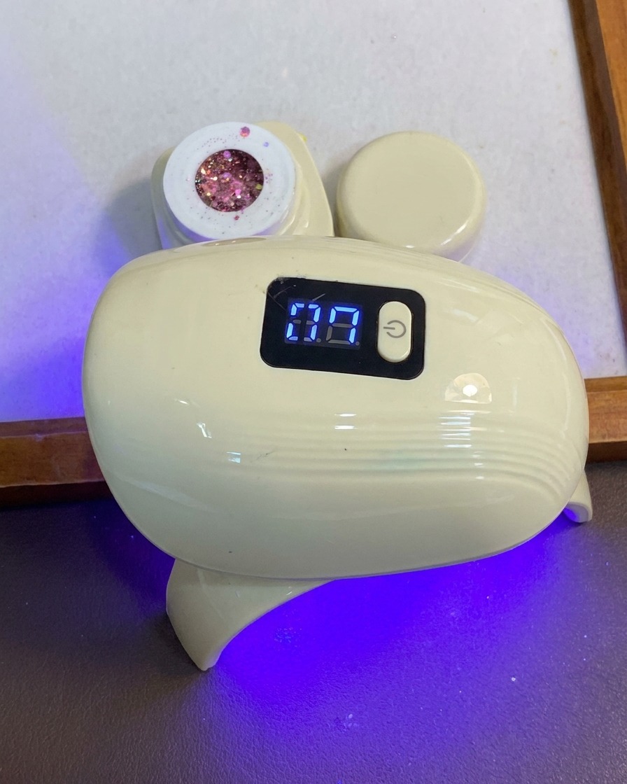 한 손이 작고 푸른 빛의 블루 앤 화이트 LED 네일 큐어링 램프 아래에 손가락을 넣고 있는 모습.
