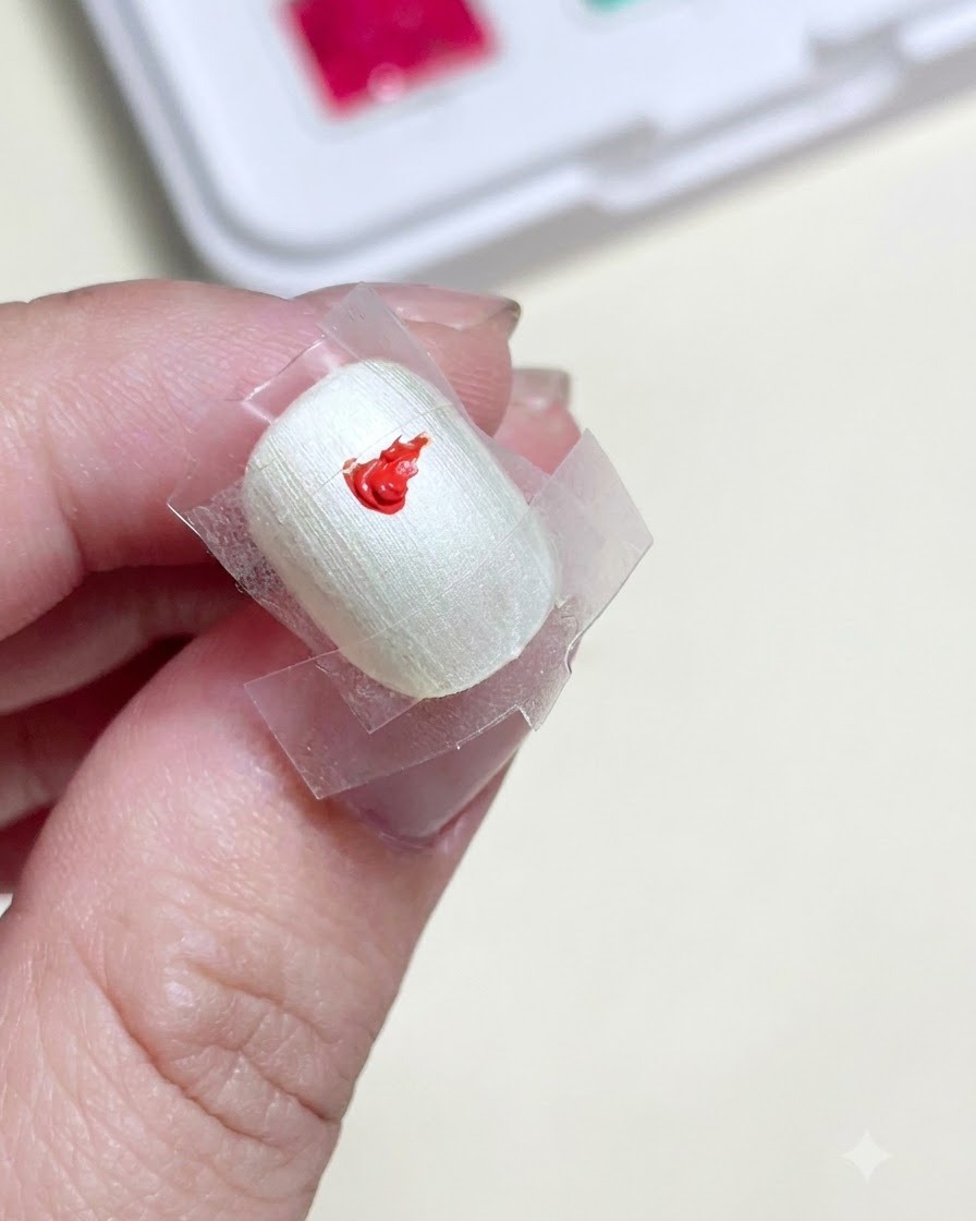 가운데에 투명 테이프 사각형이 붙여진 준비된 네일에 선명한 레드 젤 페인트 방울이 얹혀 있는 클로즈업.