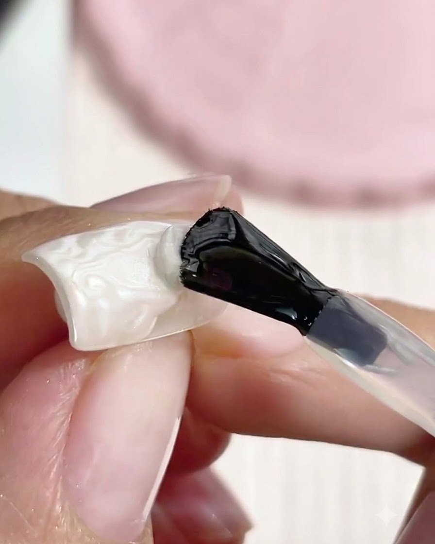 블랙 핸들 네일 브러시로 화이트 텍스처의 3D 몰딩 진주 네일 아트 피스 위에 클리어 탑코트를 바르는 모습.