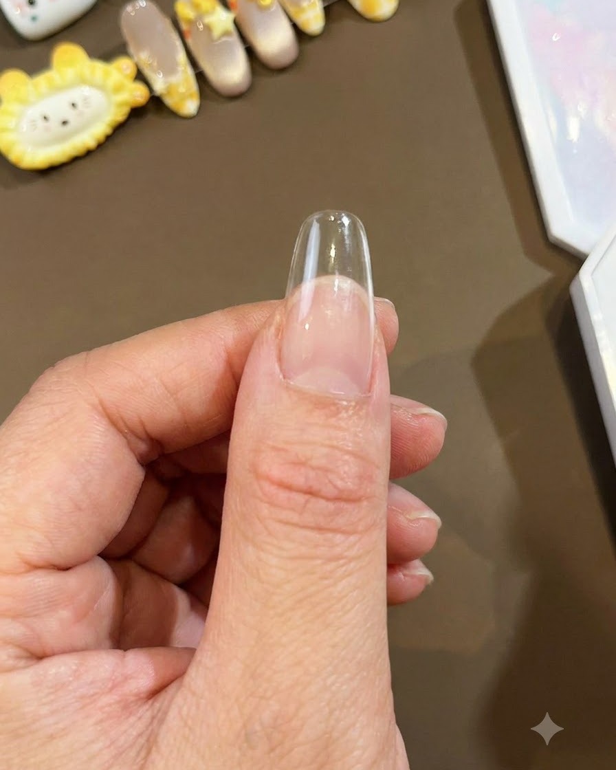 엄지손가락에 고정된 투명한 롱 아몬드 셰이프의 네일 팁으로, 아트 시술을 준비하는 모습을 담은 클로즈업 샷.