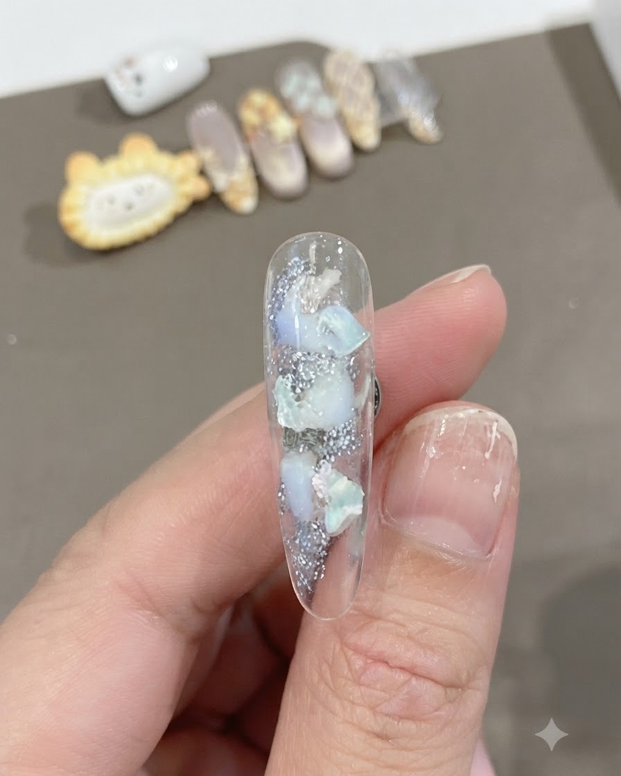 자개 조각과 반짝이는 텍스처가 내장된 투명한 롱 네일에 은은한 화이트 그라데이션 블렌드가 더해진 클로즈업 샷.