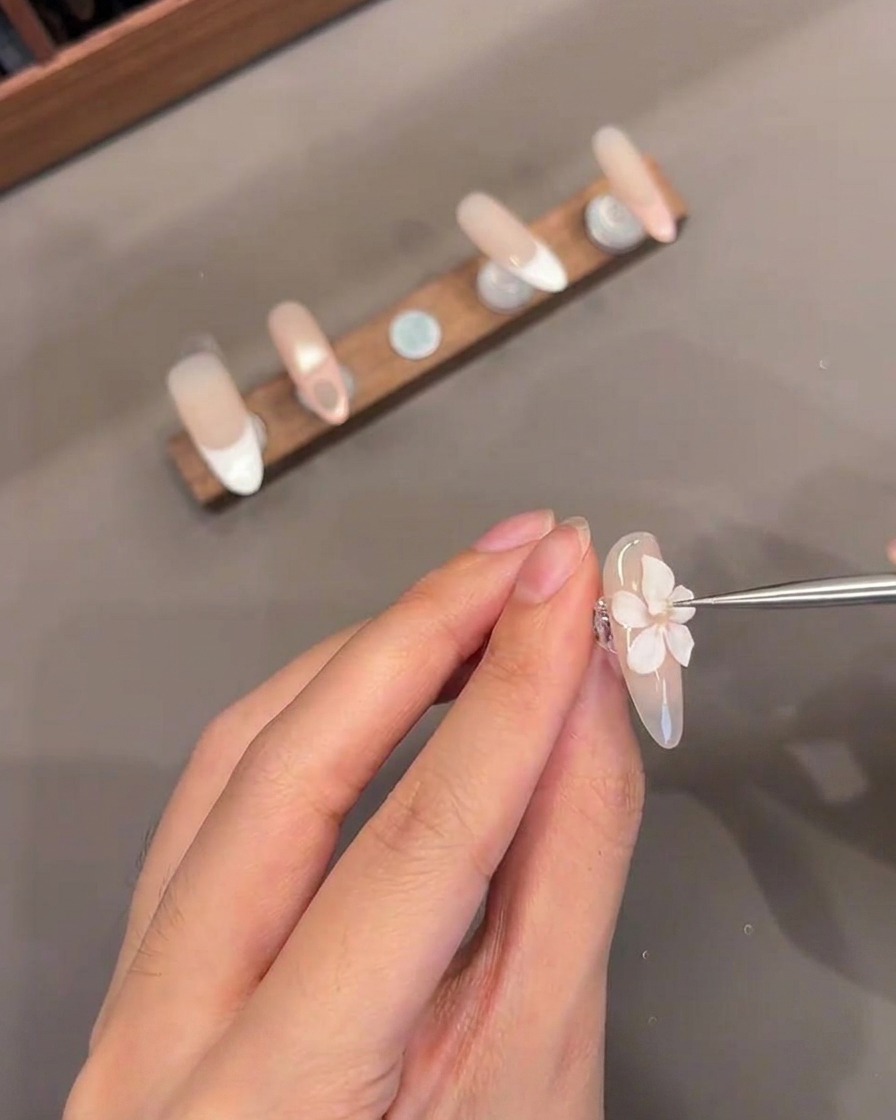 손톱 위에 조각된 3D 꽃의 중심에 얇은 도구로 작은 화이트 겔 방울을 더하는 클로즈업 샷.